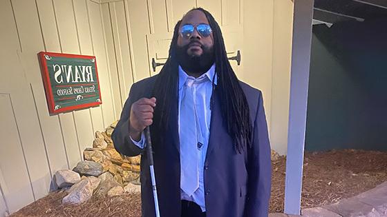 Andre Woods stands outdoors in front of a restaurant. He is wearing a blue suit and sunglasses. He is holding a mobility cane.